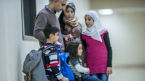 Ein Vater, eine Mutter, ein Bruder und zwei Schwestern aus dem Nahen Osten sind gerade in ihrem neuen Wohnhaus angekommen. Sie tragen ihr Gepäck bei sich. Der Vater checkt Informationen mit seinem Smartphone. Die jüngste Tochter sitzt auf dem Gepäckstück.