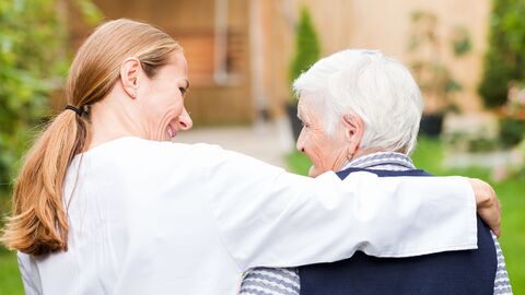 Eine junge Frau legt den Arm um eine Seniorin während sie auf ein Haus zugehen