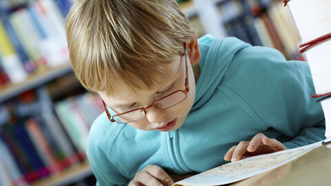 Ein Junge liest interssiert in einem Buch, im Hintergrund sind Bücherregale in einer Bibliothek