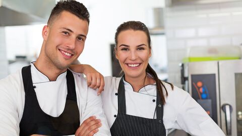 Ein junger Koch und eine junge Köchin in Arbeitskleidung in einer Küche lächeln in die Kamerat