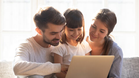 Auf einem Sofa sitzt ein Mädchen zwischen seinen Eltern und alle drei schauen auf ein Laptop, dass der Vater auf seinem Knie abstützt.