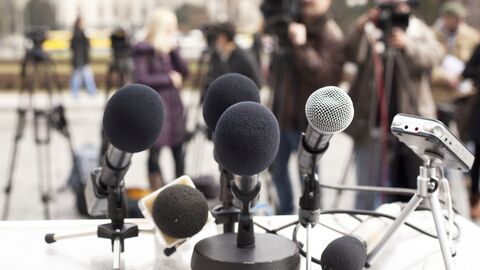 Im Vordergrund Mikrofone auf einem Tisch. Im Hintergrund, verschwommen, stehen Personen bspw. mit Schreibblock und hinter einer Kamera auf einem Stativ.