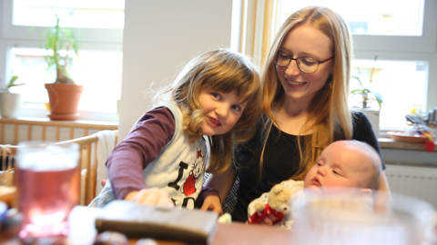 Frühe Hilfen - Ehrenamtliche Familienangebote Eine ehrenamtliche Familienhelferin hat ein Baby auf dem Arm und das Geschwisterkind neigt sich zu ihr.