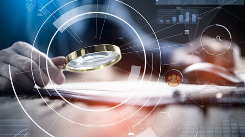 Hände mit einer Lupe und einer Computermaus auf einem Schreibtisch fotografiert