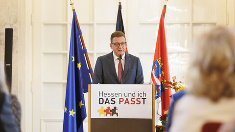 Einbürgerungsfeier 2023 Staatssekretär Stefan Sauer am Rednerpult der Einbürgerungsfeier. Im Hintergrund die Flaggen von Europa, Deutschland und Hessen.