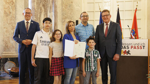 Einbürgerungsfeier 2023: Familie Elias-Samaan Minister Klose und Staatssekretär Sauer mit einer Familie. Sie stehen nebeneinander zum Gruppenfoto aufgereiht in einem Saal, im Hintergrund die hessische Flagge. Die Person in der MItte hält die Einbürgerungsurkunde in Händen.
