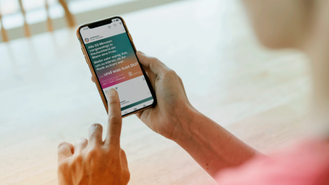Kampagnenmotiv „VERGEWALTIGUNG“ als Instagram-Beitrag Blick von hinten über die Schulter einer Frau auf ihr Handy, das sie berührt. Dort abgebildet ist das Kampagnenmotiv „VERGEWALTIGUNG“ als Instagram-Beitrag