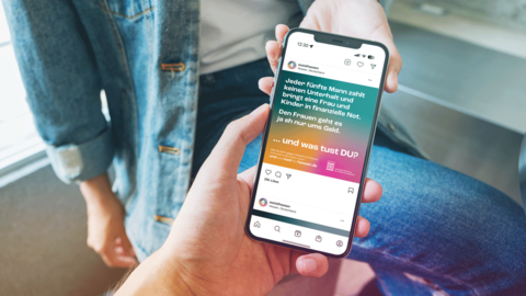Auf dem Handydisplay ist das Motiv „UNTERHALT“ Begegnungssituation: Eine weibliche Person im Hintergrund zeigt gerade einer männlichen Person im Vordergrund ihr Smartphone. Auf dem Display ist das Motiv „UNTERHALT“ zu sehen