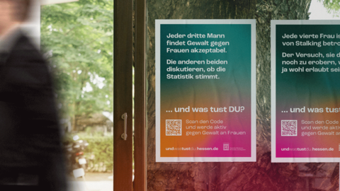 Blick von innen an eine Glastür mit zwei Plakaten. Die Motive „GEWALT GEGEN FRAUEN“ und „STALKING“ sind zu sehen. Vor der Tür geht gerade eine Person unscharf nach links aus dem Bild.