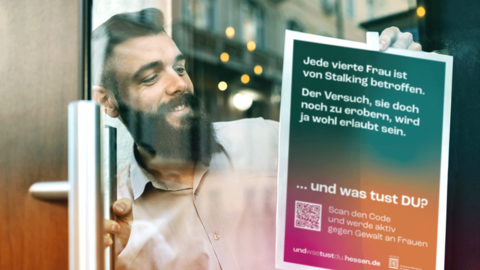 Aktionsplakat mit dem Motiv „STALKING“ Blick durch eine Glastür. Ein lächelnder bärtiger Mann bringt mit Klebeband von innen das Aktionsplakat mit dem Motiv „STALKING“ an