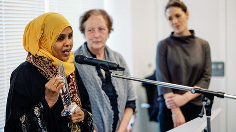 Preisträgerin Amina Sharif Mahamed Amina Sharif Mahamed steht vor einem Mikrofon und spricht, im Hintergrund sind zwei weitere Frauen zu erkennen, die ihr zugewandt sind.