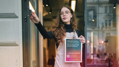 Eine junge Frau steht in der Eingangstür, sie hält ein Kampagnenposter in der Hand und macht von sich ein Selfie