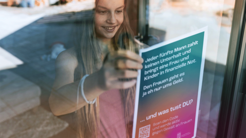 Plakat mit dem Kampagnenmotiv „UNTERHALT“ Junge Frau steht in einem Geschäft hinter der Schaufensterscheibe. Sie bringt von innen ein Plakat mit dem Kampagnenmotiv „UNTERHALT“ an