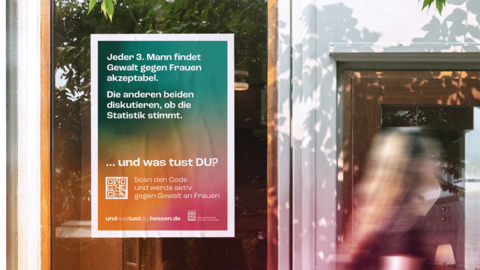 Plakat „GEWALT GEGEN FRAUEN“ Schaufensterscheibe eines Geschäfts. Hinter der Scheibe befindet sich das Plakat „GEWALT GEGEN FRAUEN“, an dem Gebäude eilt unscharf eine Passantin vorbei