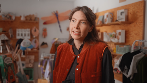 Frau in Interview-Situation- Im Hintergrund sieht man verschiedene Kleidung, Bücher und Haushaltsartikel