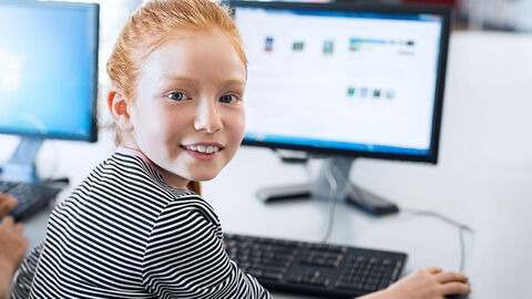 Mädchen blickt über die Schulter zum Fotografen. Sie sitzt vor einem Computer