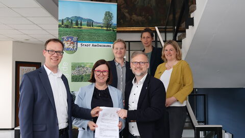 Eine Gruppe Personen rund um Staatssekretärin Strube stehen vor einem Aufsteller der Stadt Amöneburg. Sie stehen auf einer Treppe zum Gruppenfoto aufgestellt und halten ein Dokument hoch.