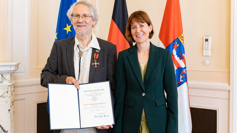 Claus Schäfer steht neben Sozialministerin Hofmann und hält seine Urkunde zum Bundesverdienstorden in den Händen, im Hintergrund die Flaggen von Europa, der Bundesrepublik Deutschland sowie Hessen.