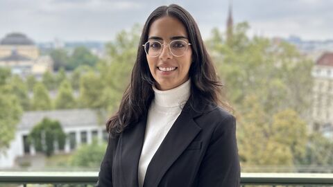 Antidiskriminierungsbeauftragte Berivan Şekerci  Frau mit dunklen Haaren mit weißem Rollkragenpullover und dunklem Blazer lächelnd vor Stadtpanorama