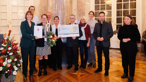 Mehrere Männer und Frauen stehen zusammen und blicken in Kamera. Teilweise heben Sie einen großen Scheck.
