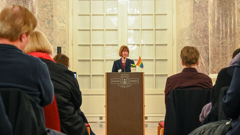 Eine Frau steht an Rednerpult. Das Publikum hört zu. 