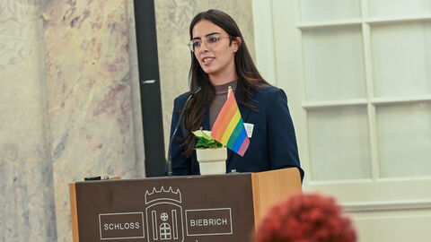 Frau steht an Rednerpult. Auf dem Pult befindet sich eine Pflanze mit einer Regenboggen-Flagge