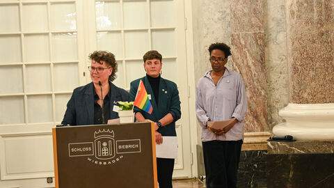 Frau steht an Rednerpult. Auf dem Pult befindet sich eine Pflanze mit einer Regenboggen-Flagge. Im Hintergrund stehen zwei weitere Frauen