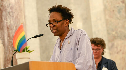 POC-Frau steht an Rednerpult. Auf dem Pult befindet sich eine Pflanze mit einer Regenboggen-Flagge
