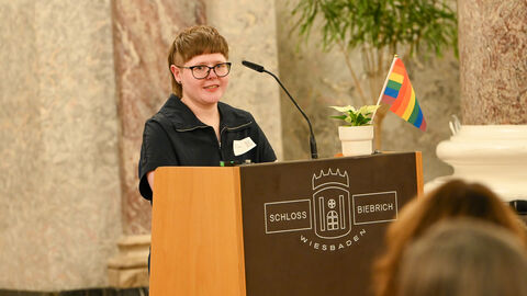 Frau steht an Rednerpult. Auf dem Pult befindet sich eine Pflanze mit einer Regenboggen-Flagge