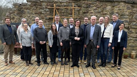 Eine Gruppe Personen steht vor einer Steinmauer in einem Außenbereich zum Gruppenfoto aufgestellt.