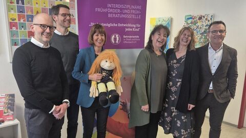 Besuch Frühförderstelle Offenbach MInisterin Hofmann steht mit weiteren Personen vor einem Aufsteller mit dem Titel "Interdiszilinäre Frühförder- und Frühberatungsstelle".