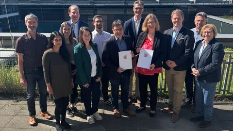 Eine Gruppe Personen steht vor einem Gebäude zum Gruppenfoto aufgestellt, vordere Personen halten Förderbescheide in den Händen.