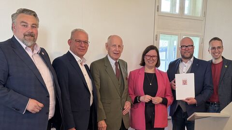 Staatssekretärin Manuela Strube steht mit fünf Männern in einer Reihe und alle lächeln. Einer hält eine Urkunde in der Hand.