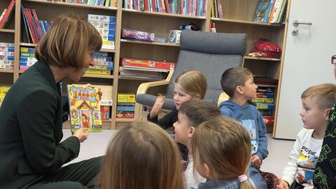 Eine Frau sitzt auf dem Boden umrundet von Kindern im Vorschulalter und hält ein Buch in den Händen. Im Hintergrund ein Bücherregal.
