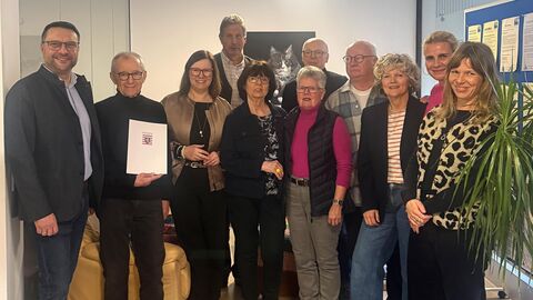 Zwölf Männer und Frauen stehen in einem Raum nebeneinander. Ein Herr auf der linken Seite hält eine Urkunde hoch.