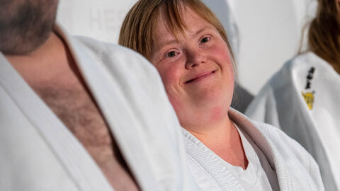 Junge Frau sitzt zwischen zwei Personen, deren Gesichter nicht zu sehen sind. Sie schaut in die Kamera und lächelt dabei.